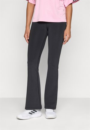 Persona che indossa leggings neri a palazzo, sneakers bianche Adidas con strisce nere e una maglietta rosa a maniche corte, in piedi su un pavimento semplice.