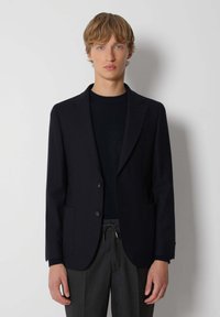 Blazer azul marino hecho de tejido texturizado con dos bolsillos frontales, cierre de un solo botón y solapas con muesca, llevado sobre un suéter negro.