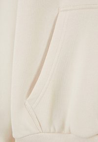 Gros plan sur un tissu beige montrant une poche avec des coutures visibles et un ourlet côtelé, typique d'un sweat-shirt ou d'un sweat à capuche.