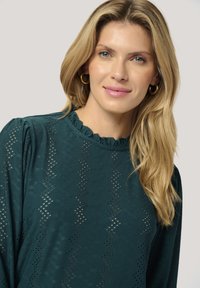 Tropfblaues Langarmshirt mit einem Rüschenausschnitt, das ein strukturiertes Muster aus Zickzack-Auschnitten und eine lockere Passform aufweist. Goldene Creolen sind sichtbar.