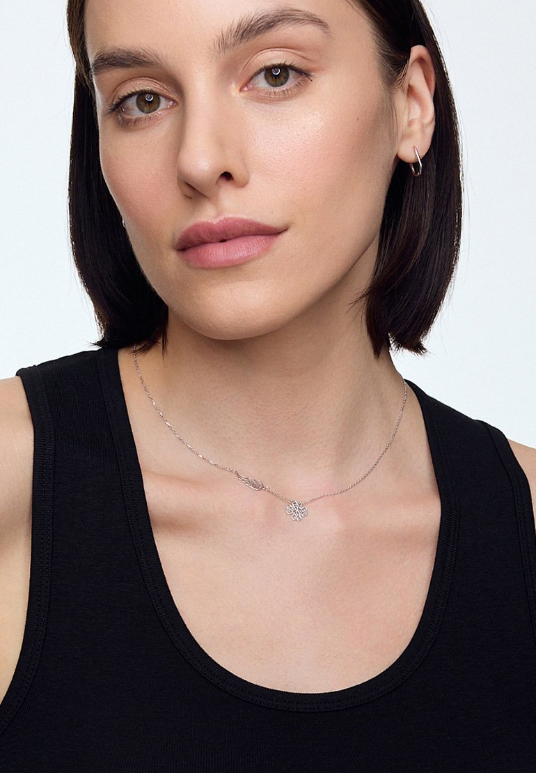 Srebrny naszyjnik z akcentem liścia i kwiatowym zawieszeniem, zaprezentowany na nagiej szyi, z czarnym topem na ramiączkach w tle.