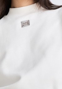 Primer plano de una persona que lleva una camiseta blanca de cuello redondo con un pequeño pin metálico cuadrado en el pecho.