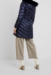 Abrigo acolchado azul marino con capucha de piel sintética, cintura ajustada y diseño de acolchado en chevron. Combinado con pantalones de pierna ancha beige y botas de tacón con estampado de serpiente blancas.