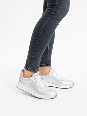 Legs wearing dark skinny jeans with silver sneakers featuring white soles and pink accents on the heels, standing on a white surface.