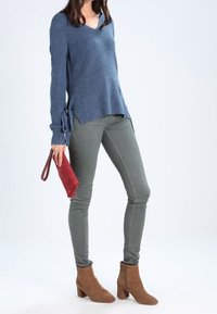 Maglione blu lavorato a maglia con scollo a V, con lacci laterali, abbinato a jeans skinny verde oliva e stivaletti marroni. Accessoriato con una borsa a tracolla rossa.
