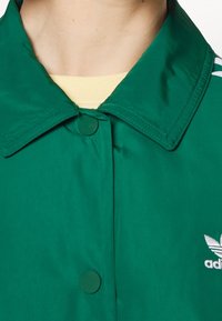 Chaqueta de poliéster verde con cierre de botones, gran cuello y acentos en blanco con tres franjas en los hombros. Pequeño logo bordado en el pecho.