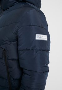 Marineblauwe donzen jas met doorgestikte structuur, ritssluiting en merkpatch op de schouder. Materiaal lijkt licht en glad.