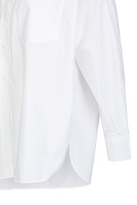 White cotton shirt featuring a button-up front, long sleeves, and a single chest pocket. The design includes a curved hem and textured fabric.