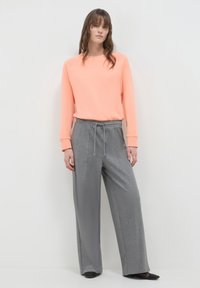 Donna in piedi contro una parete bianca, indossa una maglietta a maniche lunghe color pesche, pantaloni larghi grigi con coulisse e scarpe nere basse.