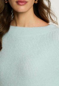 Maglione a maglia azzurro chiaro con texture a coste, scollatura ampia e tessuto morbido. Inquadratura ravvicinata con orecchini visibili.