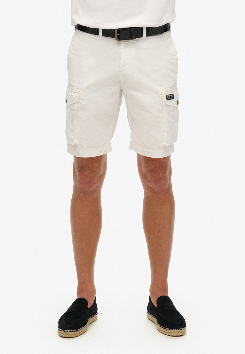 Pantalones cortos de carga blancos con dos bolsillos laterales, una textura suave y una etiqueta de parche negra en el bolsillo. Combinados con mocasines negros.