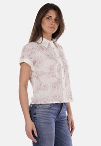 Maglietta a maniche corte in tessuto bianco con un motivo floreale rosa, bottoni frontali e colletto. Abbinata a jeans blu.