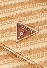 Borsa intrecciata in paglia beige con un logo triangolare in bronzo "GUESS USA" che presenta un punto interrogativo, evidenziando l'artigianato e il design testurizzato.
