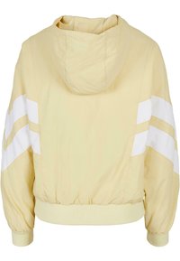 Ljusgul hoodiejacka med vita ränder på ärmarna, elastiska manschetter och cinchad nederkant. Tillverkad av lätt, texturerat tyg.