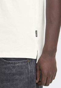 Camiseta de algodón en color blanco roto con una abertura lateral, que presenta una etiqueta negra "BLEND" en el dobladillo, combinada con jeans de mezclilla gris.