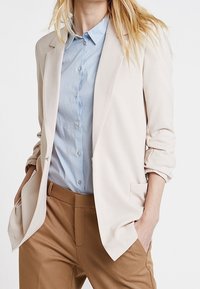 Béžový blazer s klopami a dvěma předními kapsami, spárovaný se světle modrou košilí s knoflíky a béžovými kalhotami. Měkká textura látky.