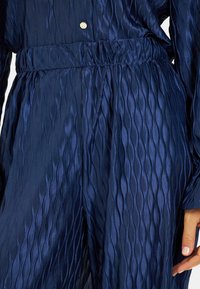 Granatowy, teksturowany materiał z falistymi plisami, z gromadzonym w pasie i dekoracyjnym złotym guzikiem przy dekolcie.