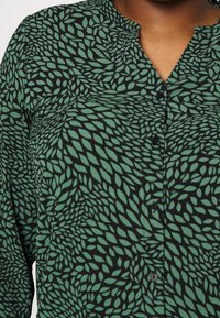 Camicia nera con un motivo a foglia verde, parte anteriore con bottoni e colletto alla coreana. Realizzata in tessuto leggero con una texture liscia.