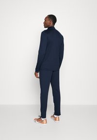 Marineblauw sportief tweedelig outfit met een langemouwen top en bijpassende broek, gecombineerd met bronzen en witte sneakers. Zacht materiaal en een strakke pasvorm.