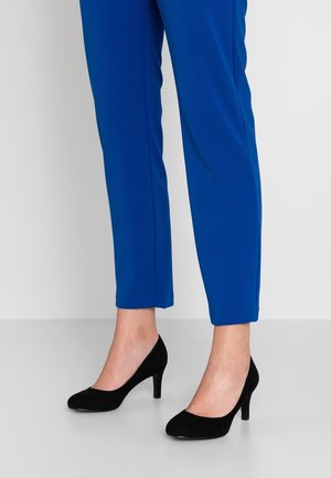 Beine in leuchtend blauen Slim-Fit-Hosen kombiniert mit schwarzen Wildleder-High-Heels vor schlichtem hellem Hintergrund.