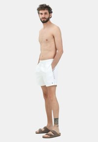 Costumi da bagno bianchi con vita regolabile tramite cordoncino, tasche laterali e logo sul gastro sinistro. Abbinati a sandali marroni.