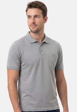 Mężczyzna w jasnoszarej koszulce polo z krótkim rękawem i przednią kieszenią, patrzący lekko w prawo na jednolitym białym tle.