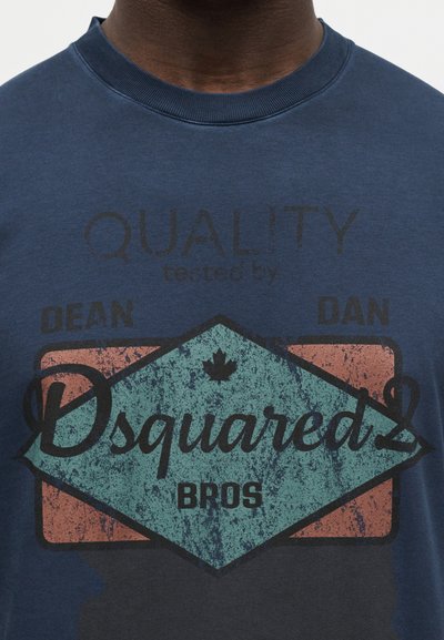 Μπλε ναυτικό πουκάμισο με φθαρμένο κείμενο «Quality tested by Dean Dan Dsquared2 Bros» και ένα μικρό φύλλο σφενδάμου πάνω από το όνομα της μάρκας.