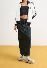 Persona che indossa una giacca bianca Adidas, un top corto nero, una gonna lunga a righe nere, calzini bianchi e zoccoli trasparenti con piattaforma su un pavimento giallo.