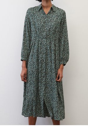 Kobieta w długiej sukience midi z długim rękawem i zapięciem na guziki, z zielono-czarnym kwiatowym wzorem, stojąca na tle jednolitej jasnej ściany.