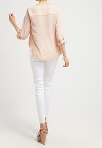 Vaaleanpunainen, puoliksi läpinäkyvä paita, jossa on käärityt hihat ja pyöristetty helma. Yhdistetty valkoisiin istuviin housuihin ja nude-väriin loivissa korkokengissä.