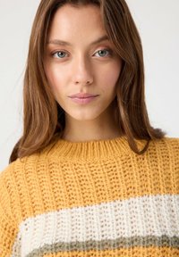 Suéter de punto en amarillo mostaza con textura gruesa, que presenta franjas horizontales blancas y verdes olivo cerca del dobladillo. Cuello redondo.