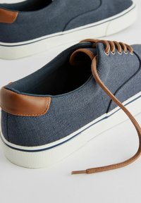 Marineblauwe canvas sneakers met bruin leren accenten, witte rubberen zool en platte veters; textielstof en contrasterende stiksels.