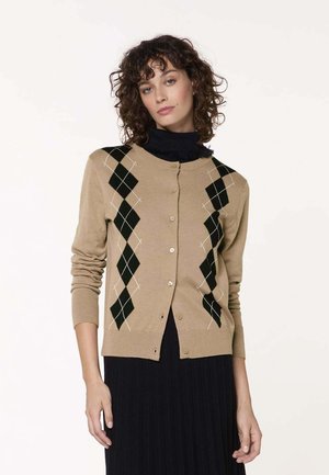 Frau mit lockigem Haar, die eine beige, mit Knöpfen versehene Strickjacke mit schwarzem Rautenmuster über einem schwarzen Rollkragenpullover und schwarzem Rock trägt, vor weißem Hintergrund stehend.