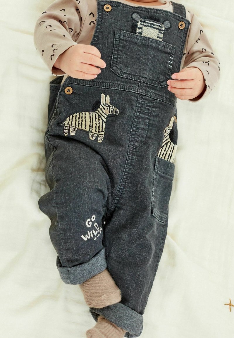 Bébé portant une salopette foncée avec des patches zèbre et le texte "Go Wild!", associée à une chemise beige à manches longues et des chaussettes marron clair.