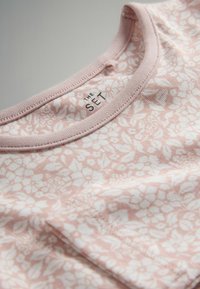 Bledoružový top s kvetinovým vzorom, okrúhlym výstrihom, prúžkovaným lemovaním a malou nálepkou s nápisom "THE SET" pri golieri. Mäkka textúra látky.