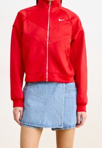 Röd zip-up jacka med hög krage och chevron-sömmar, gjord av slät tyg, ihop med en blå denimkjol med fickor.