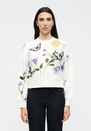 Γυναίκα με μακριά σκούρα μαλλιά που φοράει ένα λευκό φούτερ διακοσμημένο με κεντητά λουλούδια και μπλε πεταλούδες, σε συνδυασμό με σκούρα τζιν.