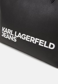 Črna teksturirana torba z bold belim napisom "KARL LAGERFELD JEANS". Oblikovana je pravokotno s gladkimi robovi in minimalističnim estetskim pristopom.