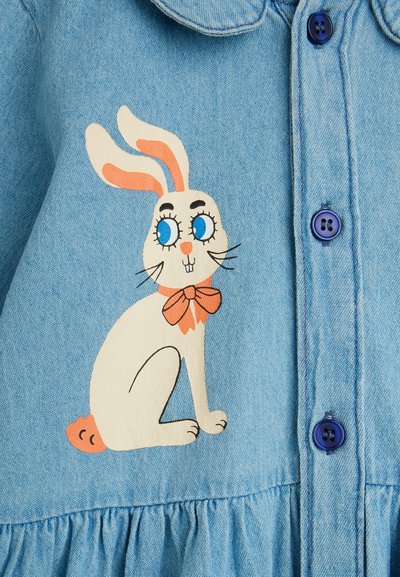 Lapin blanc de dessin animé aux yeux bleus, oreilles intérieures orange et nœud sur un tissu bleu clair boutonné.