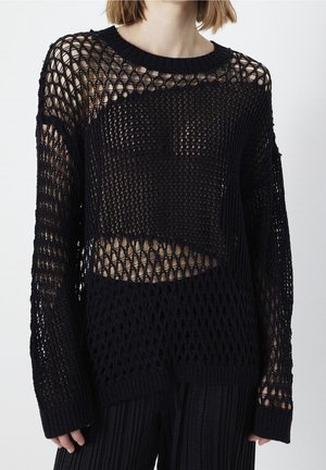 Femme portant un pull noir à manches longues en maille ajourée sur un top noir, associé à un pantalon noir texturé, sur un fond clair uni.