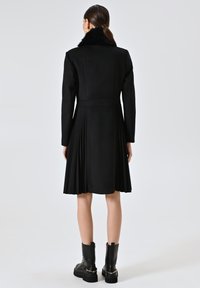 Schwarzer Wollmantel mit Faltenrockdesign, ausgestattet mit einem Pelzkragen, langen Ärmeln und knielanger Länge, kombiniert mit schwarzen Stiefeletten.
