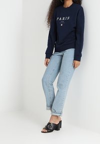 Sudadera azul marino con el texto "PARIS", jeans rectos azul claro y sandalias negras de tacón con un acento circular.