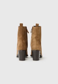Bottines en suede marron avec un talon épais et des détails en zip, vues de l'arrière sur un fond gris clair.