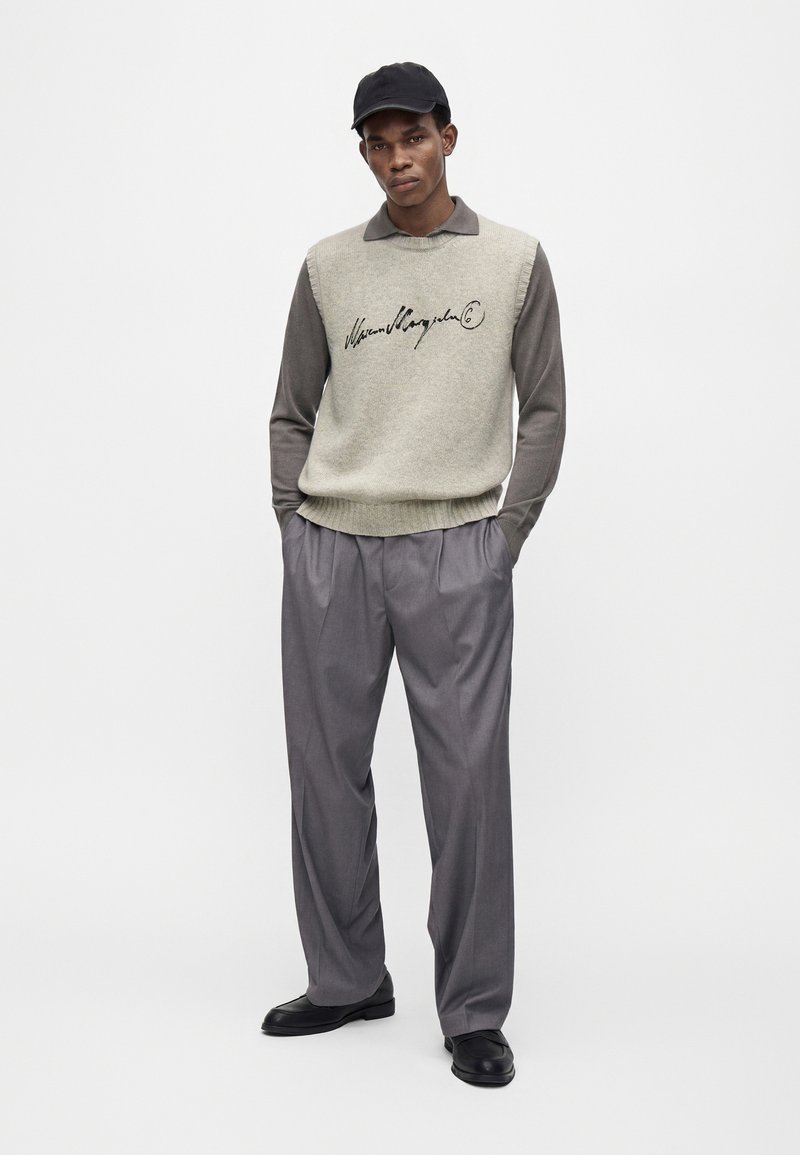 Mann trägt schwarze Kappe, hellgrauen ärmellosen Pullover mit schwarzer Schrift, dunkelgraues Langarmhemd, lockere graue Hose und schwarze Schuhe, steht mit Händen in den Taschen.