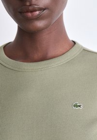 Olivgrünes, geripptes Baumwoll-T-Shirt mit rundem Halsausschnitt und kleinem gesticktem Krokodil-Logo auf der Brust. Glatte Textur.