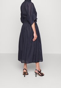 Nainen, jolla on päällä tummansininen midi-mekko, jossa on kolme neljännesosaa hihat, ja mustat nauhaketjut korkokengät, seisoo yksinkertaisen taustan edessä.