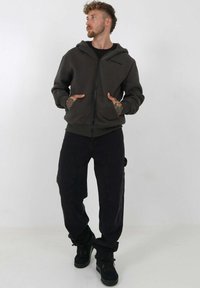 Donkergroene zip hoodie met een kenguruzak, gecombineerd met zwarte cargobroeken. Gelaagd stof en casual pasvorm met zichtbare stikdetails.