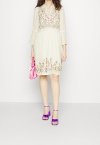 Krämdress med blommig broderi, trumpetärmar och midjeband. Matchad med lila klacksandaler och en rosa handväska.