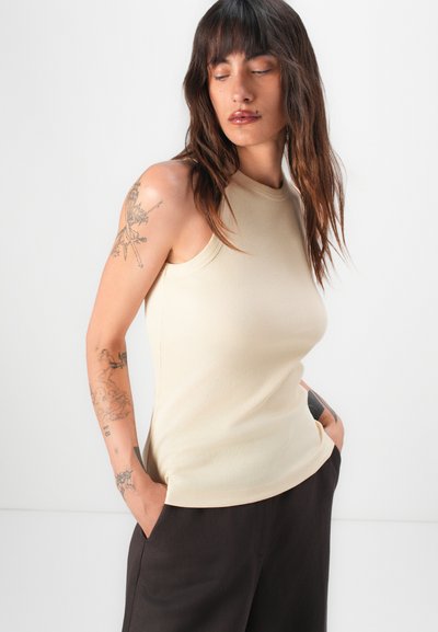 Top bej din bumbac cu dungi, fără mâneci, cu guler înalt, textură moale și croi ajustat, asortat cu pantaloni negri tip evazat.