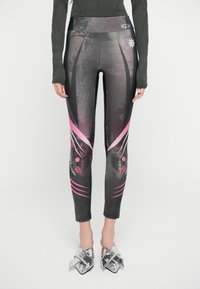Kvinne iført svarte og rosa mønstrede leggings, metalliske spisse sko med store sløyfer, og en mørk langermet topp med tommelhull.
